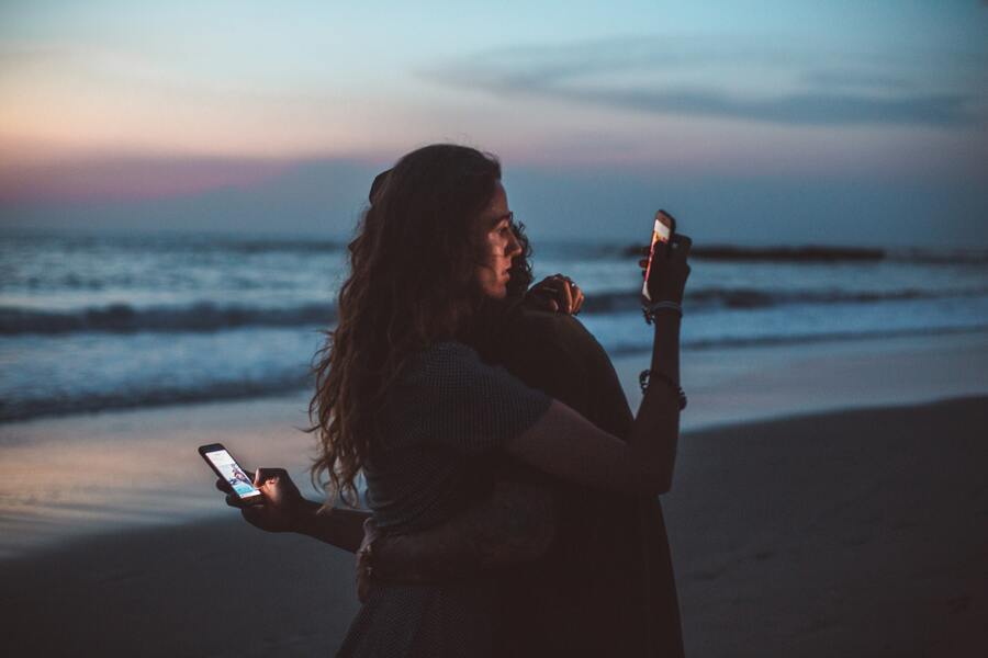 casal se abraçando em uma praia e cada um olhando para o celular pelas costas um do outro
