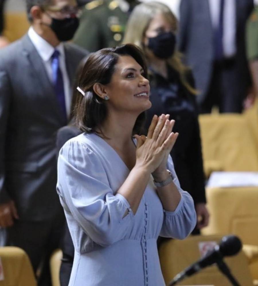 Foto de Michelle Bolsonaro em cerimônia na Escola Superior de Defesa. Ela está de pé, sorridente e batendo as palmas da mão. Ela usa vestido azul.