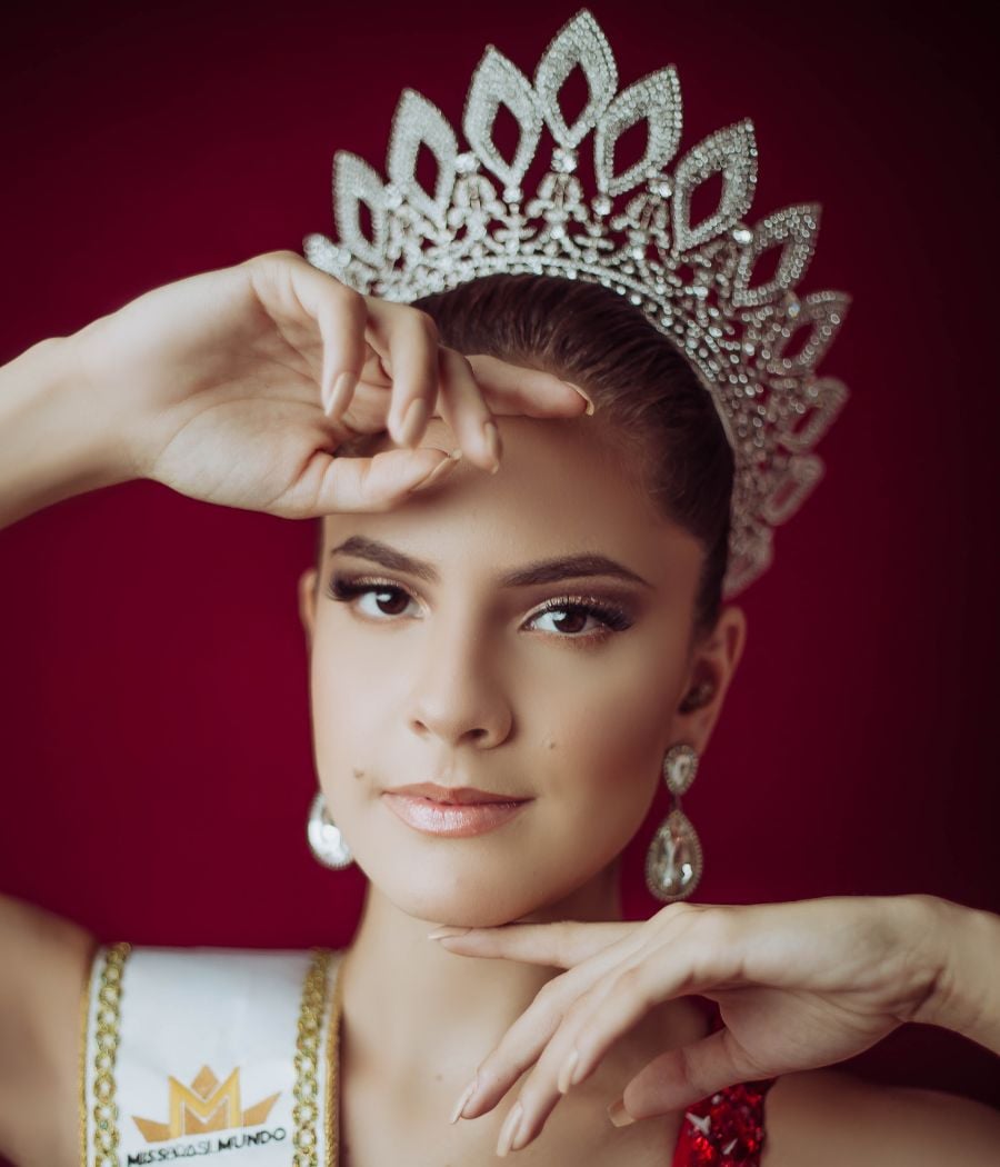 A candidata de Pernambuco ao Miss Brasil Mundo posa em frente a um fundo vermelho com uma coroa