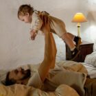 pai deitado na cama segurando um bebê loiro acima de seu corpo