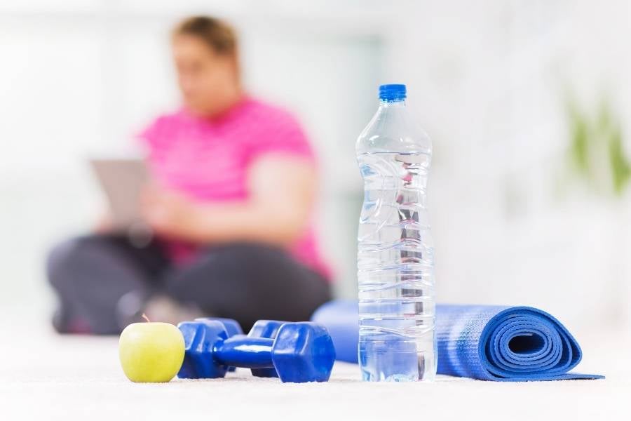 Mulher sedentária ao lado de garrafa de água, pesos, maçã e tapete de yoga