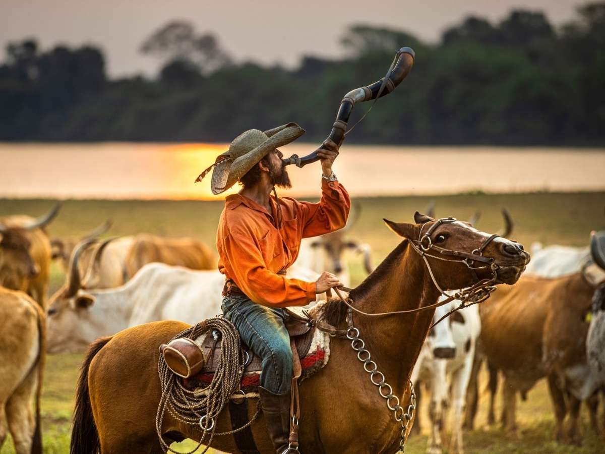 Foto de José Leôncio (Renato Goés) em cavalo tocando boiada com som de berrante. Ao fundo, paisagem do pantanal, lago e árvores.