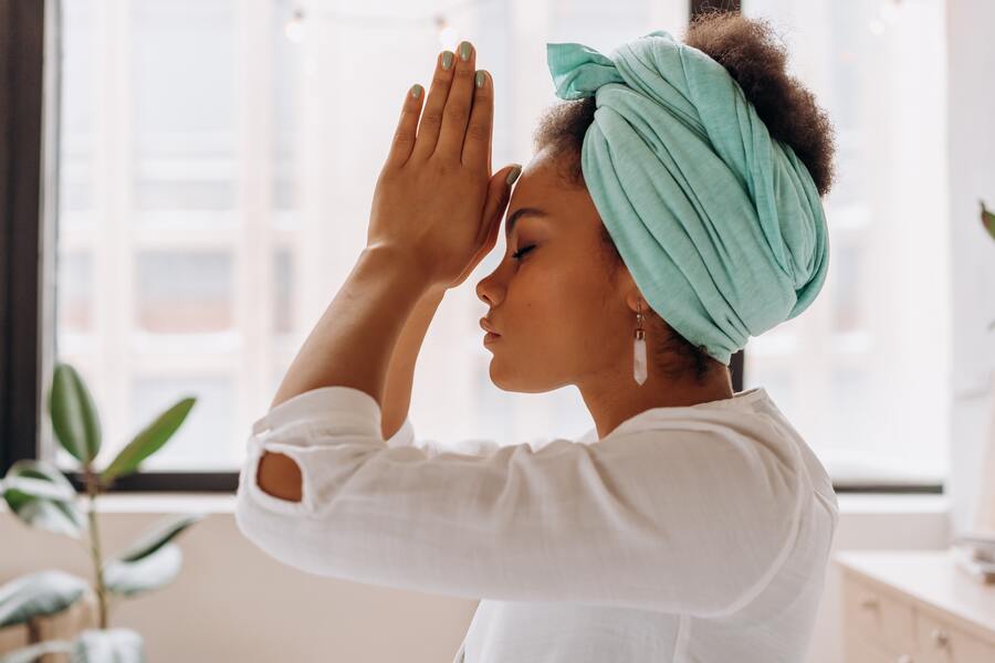 mulher negra com um lenço azul claro na cabeça colocando as mãoes em cima da teste em formato de oração para aplicação de reiki no chakra frontal