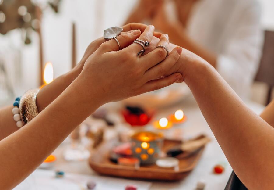 duas mãos femininas com diversos anéis e uma mesa com cristais e velas