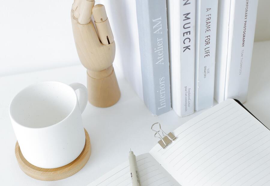 mesa organizada com uma xícara branca, livros, uma mão de madeira e um caderno com pauta em branco