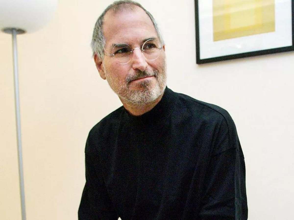 Foto de Steve Jobs da cabeça à cintura. Fotografado em sala com luminária e quadro na parede.