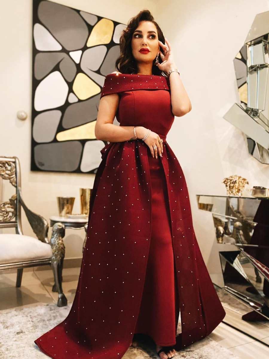 Cantora árabe Amal Al Anbary usando vestido na cor vinho da AAVVA. Ela está em pé em um local com decoração luxuosa de cadeiras, quadros e espelho.