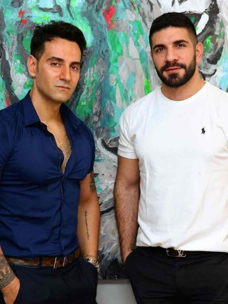 Vincenzo Visciglia e Ahmad Ammar de pé com as mãos no bolso posando para foto de frente a quadro com pintura abstrata.