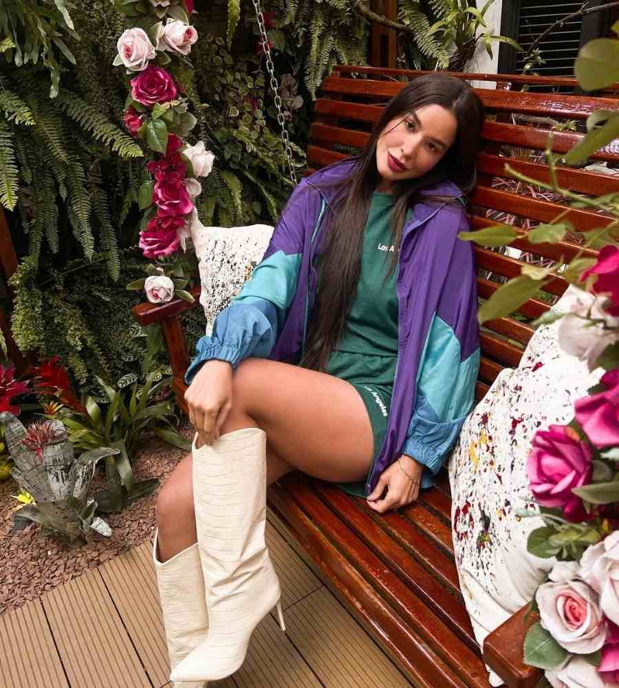 Foto da ex-bbb sentada em balanço de madeira com almofadas e flores ao redor. Ela usa conjunto de blusa e shorts confortáveis verdes, com blusa de frio roxa e azul e bota de cano alta off white.