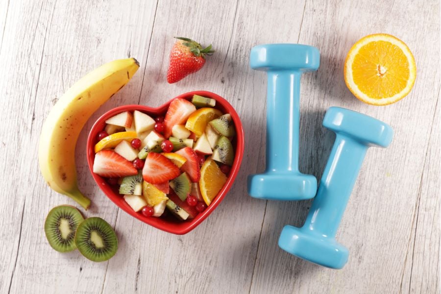 foto de salada de frutas em tigela de coração ao lado de halteres azuis, simbolizando exercício físico e alimentação saudável