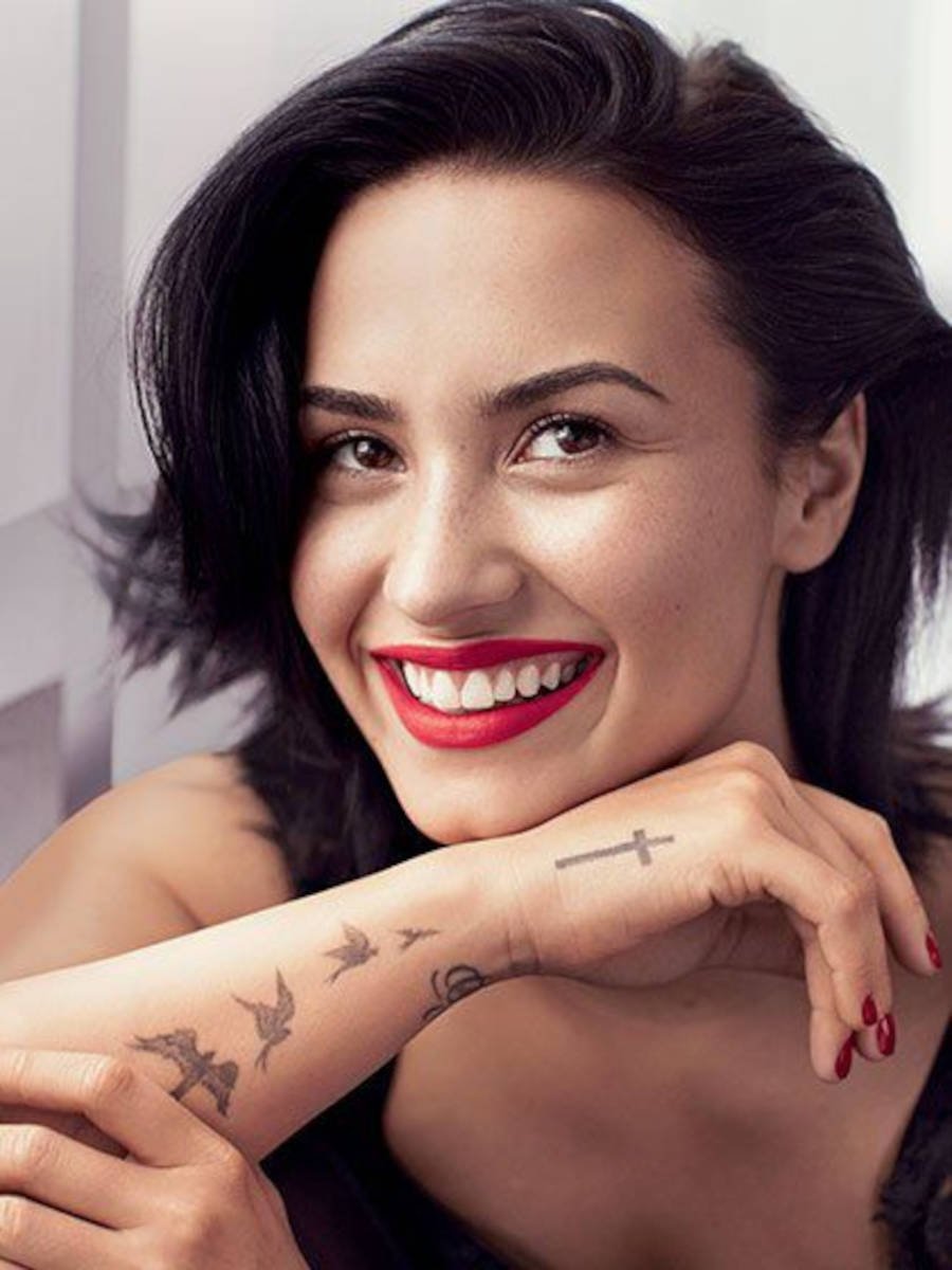 Foto de Demi sorrindo de batom vermelho com a tatuagem de cruz na mão