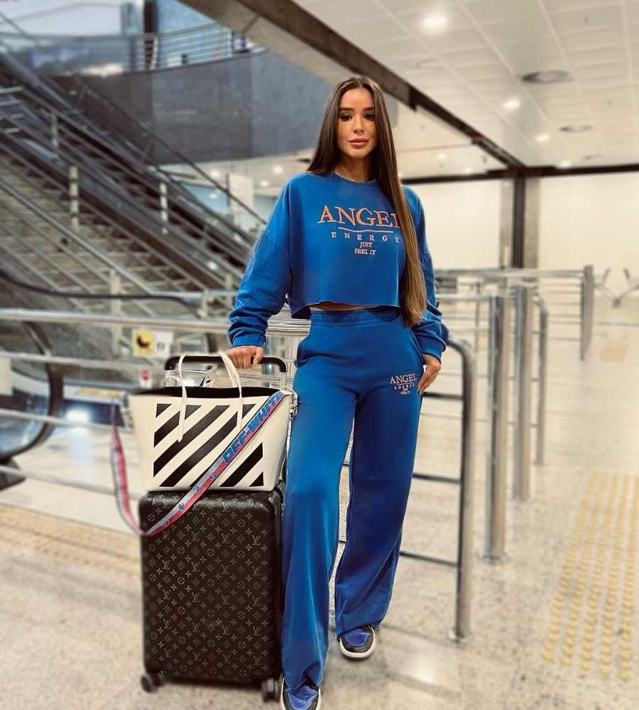 Foto de Kerline com conjunto de frio moletom azul. Ela apoia uma das mãos em mala de rodinha com bolsa em cima. Está em aeroporto ao lado das escadas rolantes.