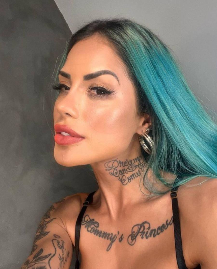 Imagem em fundo cinza-claro e cinza-escuro. No centro selfie de Tati Zaqui usando roupa de alcinha preta sorrindo.  Ela tem o cabelo azul e tatuagens espalhadas pelo corpo todo.