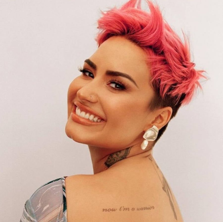 Foto de Demi Lovato  de costas com o cabelo curto rosa e com a tatuagem "Now i'm a warrior" em destaque