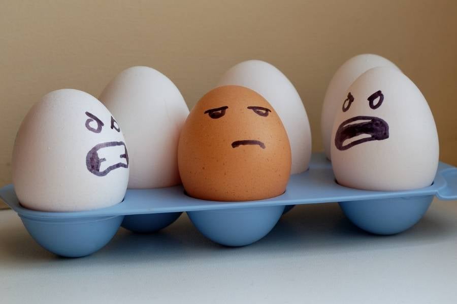 Ovos brancos pintados com cara de raiva olhando para um ovo marrom com cara de triste