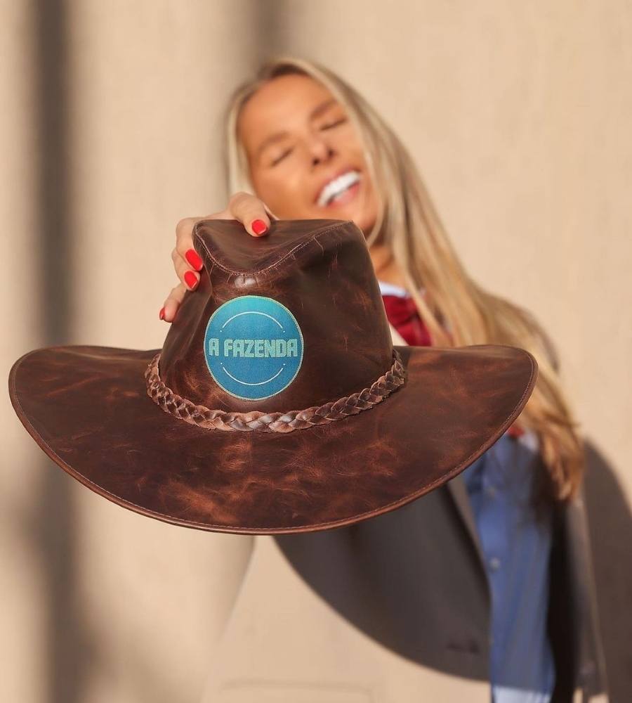 Foto de Adriane Galisteu sorridente segurando cahepéu de couro com a logotipo do A Fazenda.