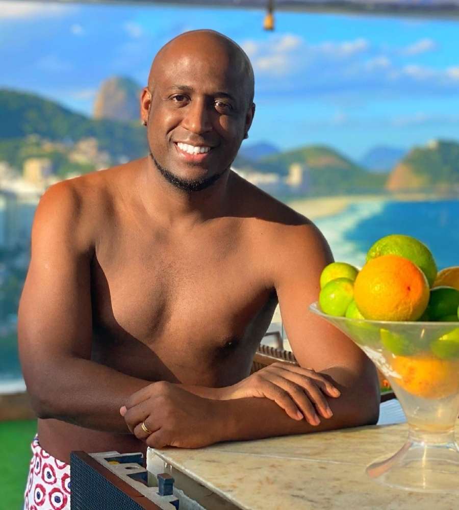 Foto de André Marinho encostado em bancada de pedra com pote cheio de laranjas e limões. Ele está de sunga e em praia.