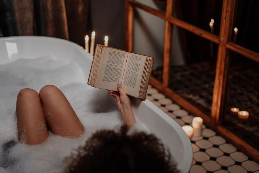 mulher em uma banheira com espuma lendo um livro com velas no chão