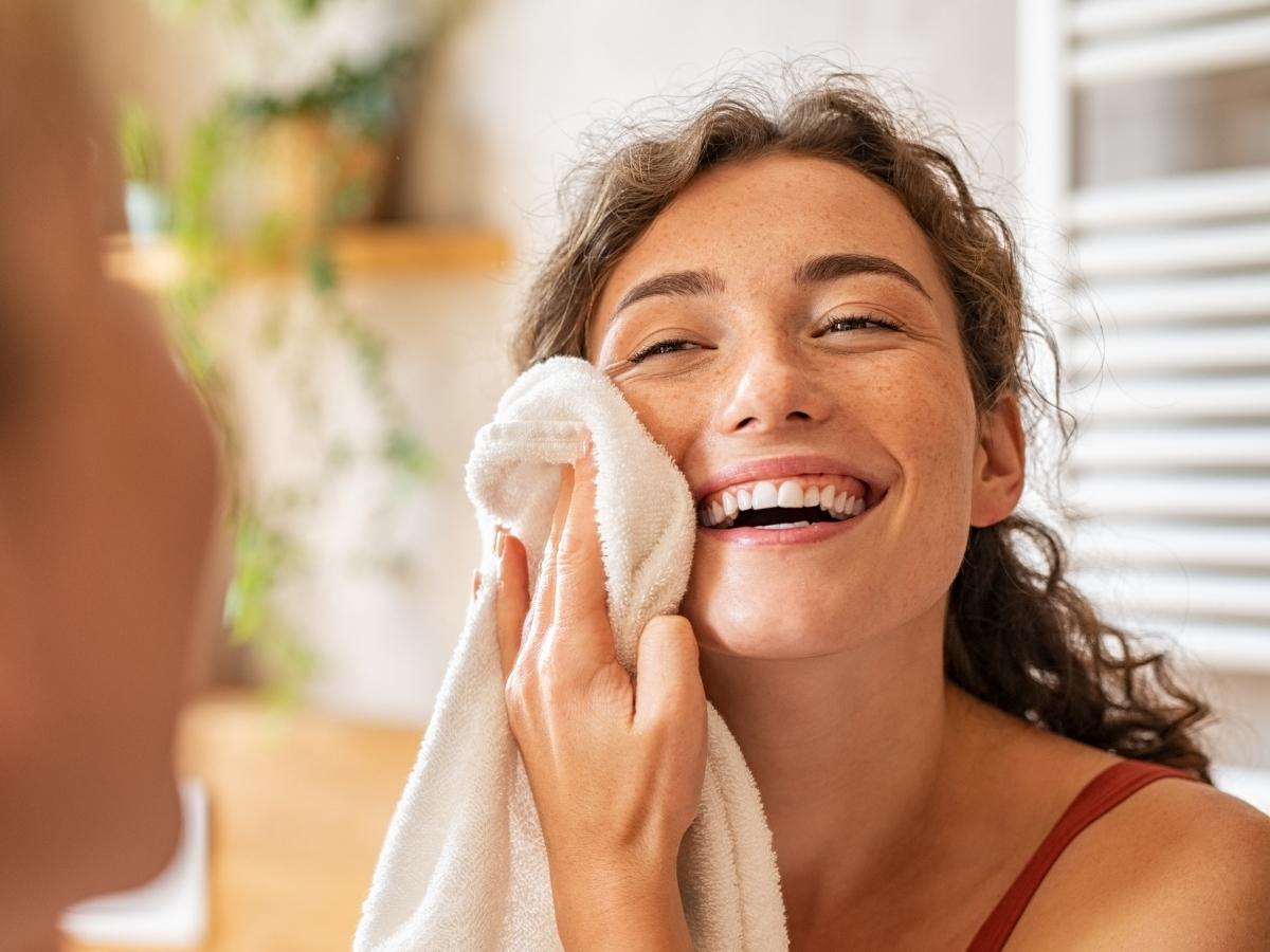 Foto de mulher sorridente secando o rosto com toalha branca em frente ao espelho.