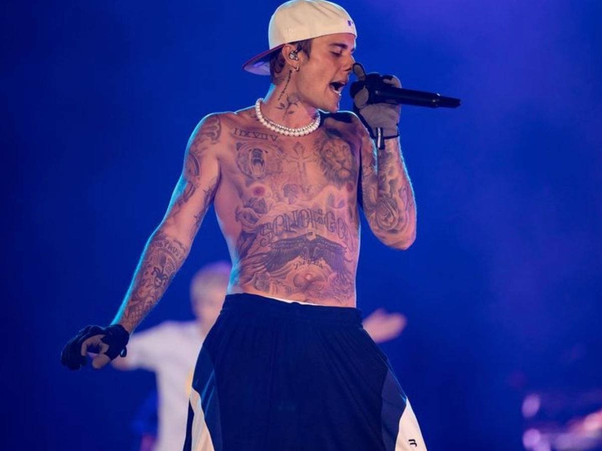 Foto do cantor Justin Bieber sem camisa e cantando em um palco com o fundo azul