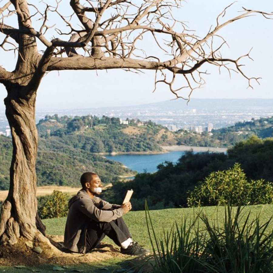 Ator do filme As Mil Palavras sentado debaixo da árvore com a boca tapada e lendo um livro. 