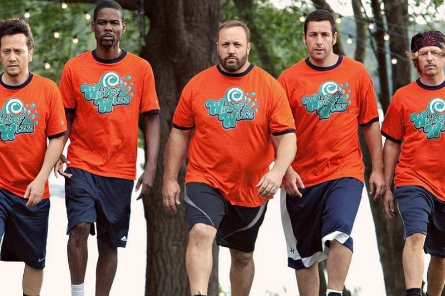 Foto de 5 homens atores do filme "Gente Grande" usando roupas iguais: camiseta laranja e shorts preto. Eles estão andando de frente para a câmera e ao fundo tem árvores com luzes.