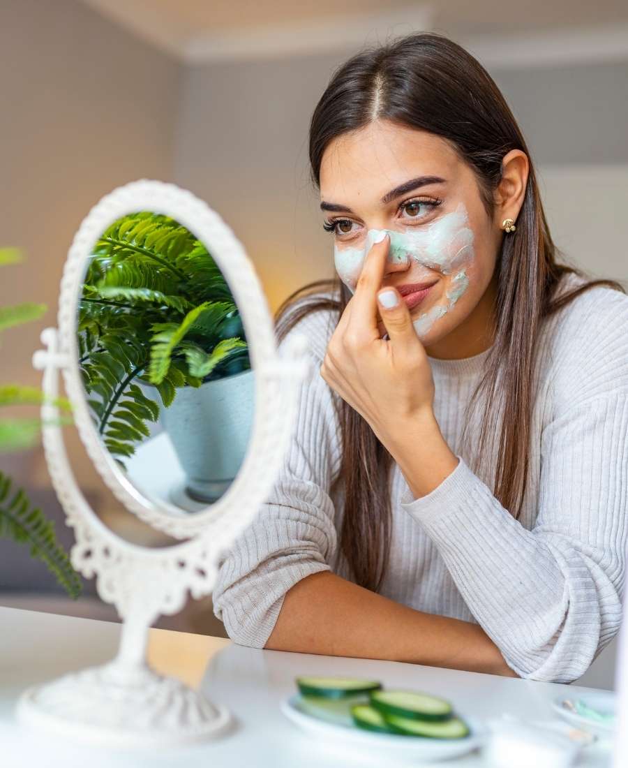 Foto de mulher sentada olhando para um espelho de mesa. Ela passa creme no rosto.