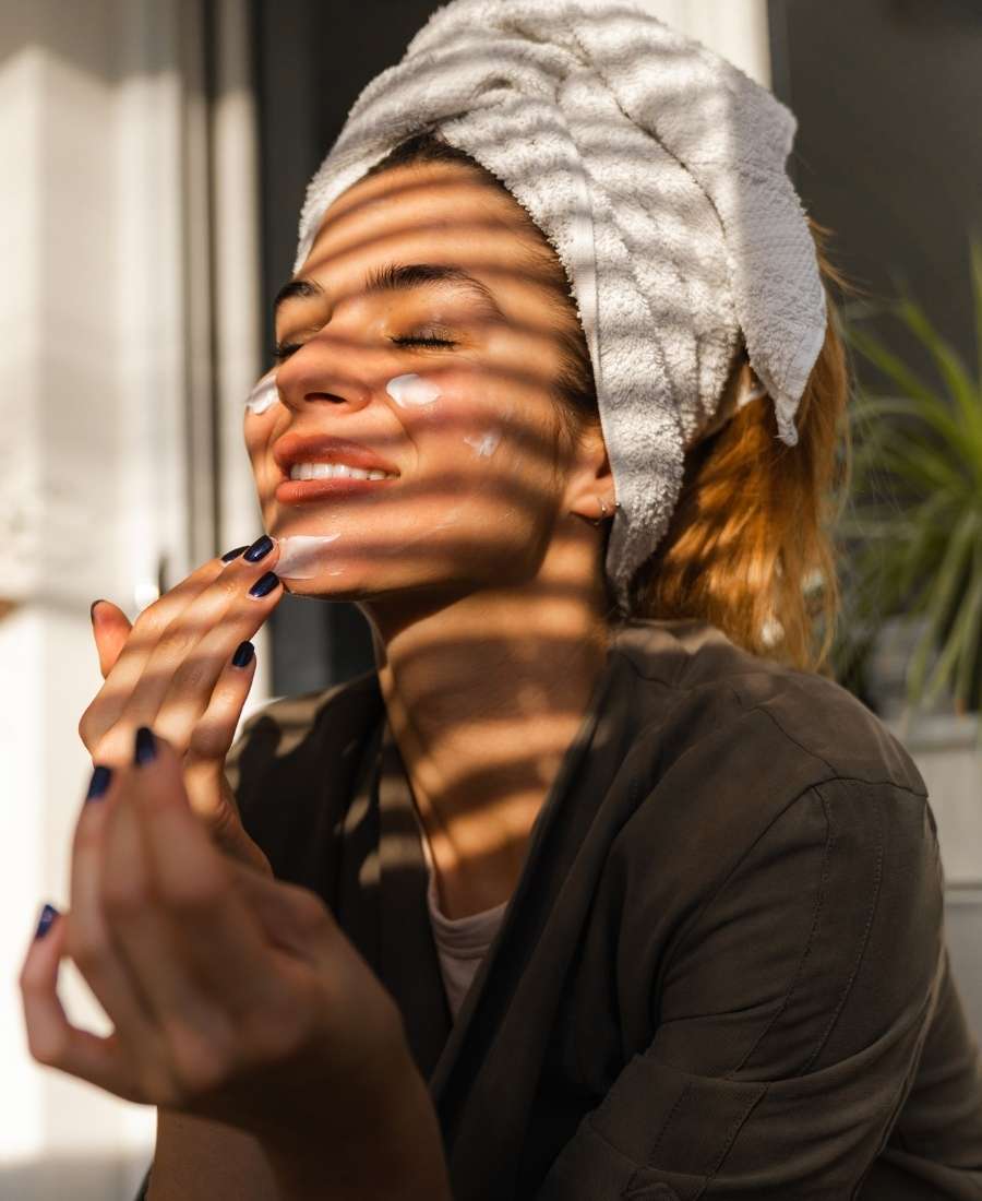 Foto de mulher com os olhos fechados passando creme no rosto e usando toalha no cabelo.