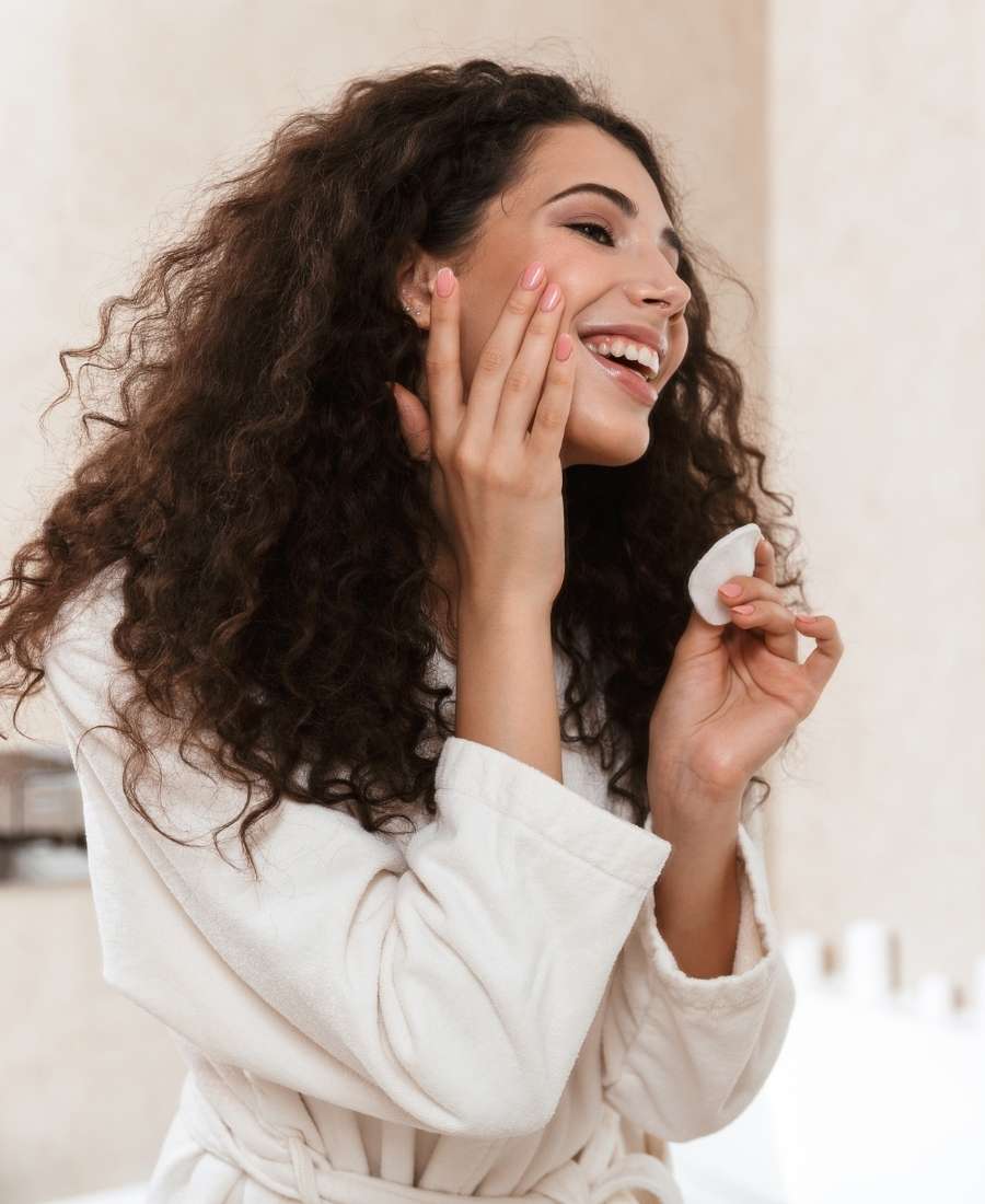 Foto de mulher sorridente com a mão no rosto e outra segurando disco de algodão. 