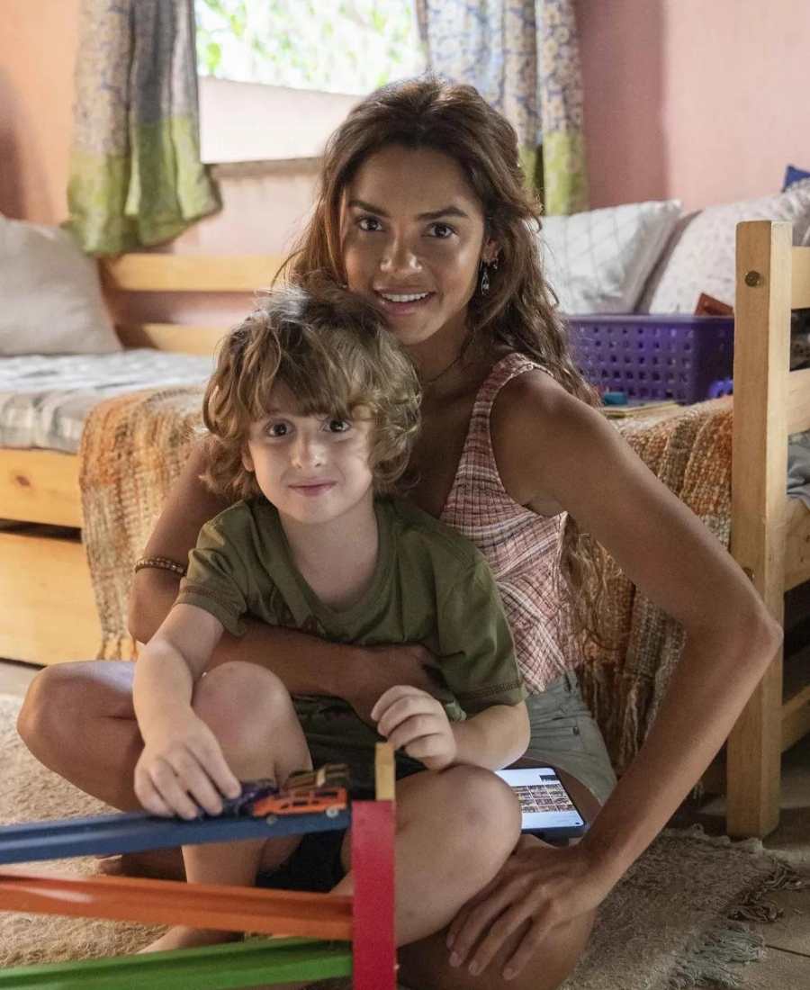 Foto de Lucy Alves como Brisa e seu filho na novela. Os dois estão sentados no tapete ao lado da cama.