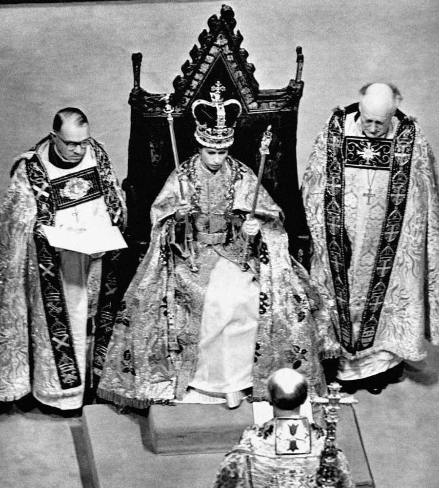 Foto da Rainha Elizabeth II no dia de sua coroação. Está sentada em seu trono ao lado de padres.