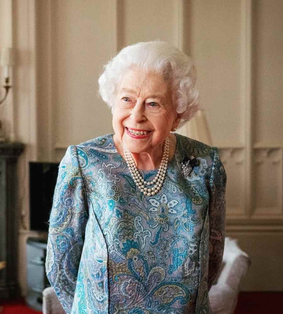 Foto da monarca britânica hoje em dia, está usando vestido floral azul, colares de pérolas e está de pé em sala com poltronas.