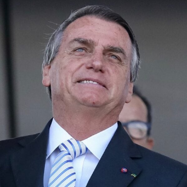 Foto de Jair Bolsonaro sorrindo, usando um terno preto, camisa branca e gravata azul e branca