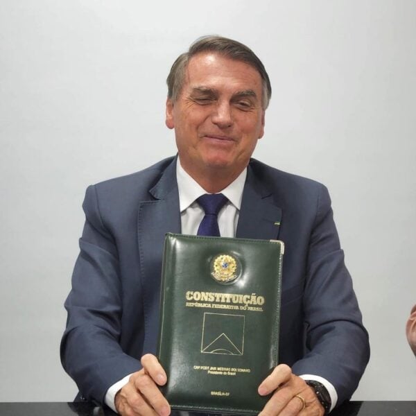 Foto de Jair Bolsonaro sentado, de terno, sorrindo e segurando a Constituição Brasileira nas mãos