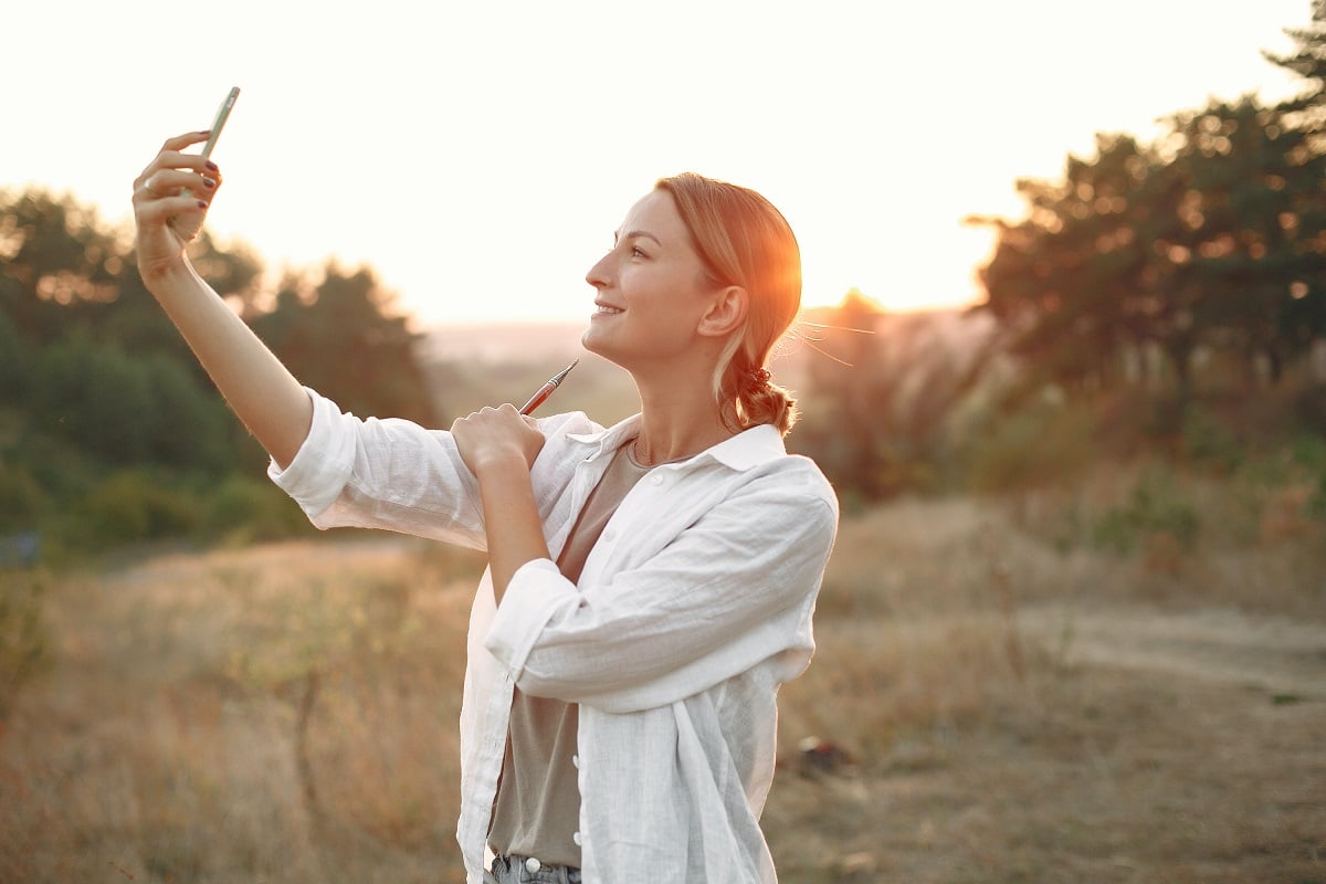 Mulher de casaco branco tirando uma selfie sorrindo no meio da natureza
