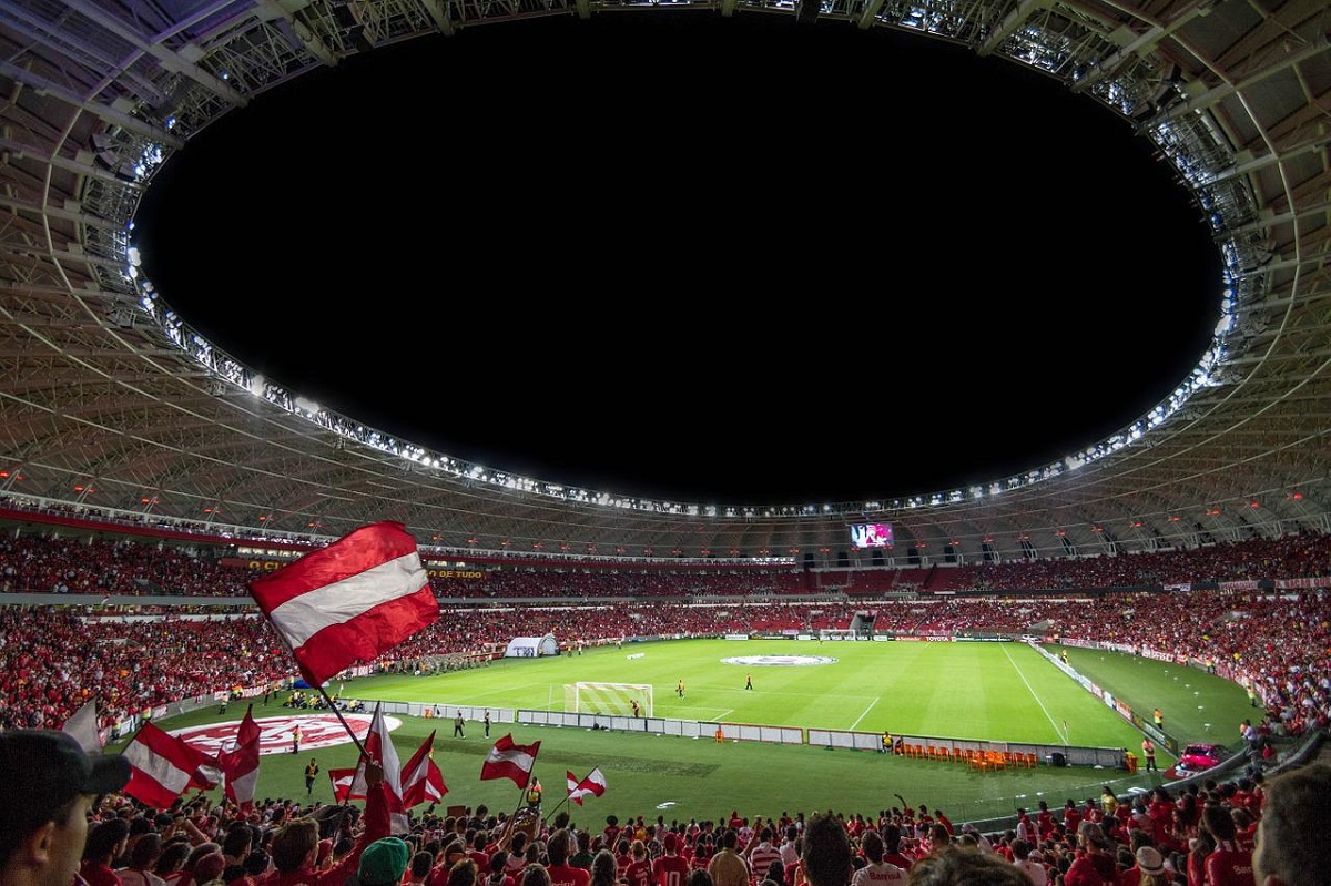 Foto mostra um estádio todo iluminado e repleto de pessoas levantando bandeiras vermelhas com branco e assistindo a uma partida noturna