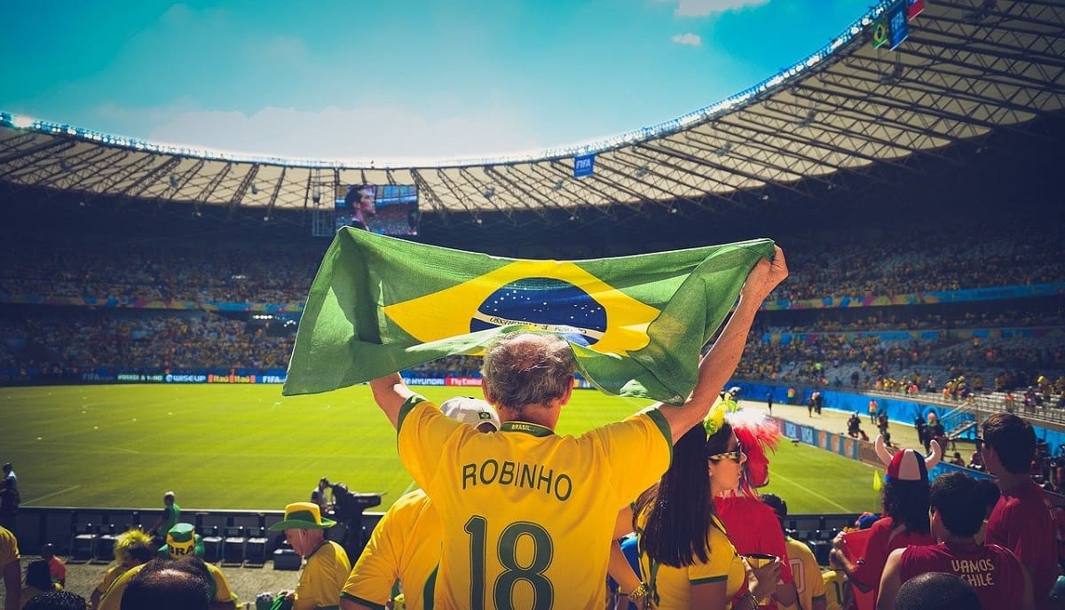 Foto de um idoso usando a blusa do Brasil com o nome de Robinho e segurando uma bandeira do Brasil. Ele está em um estádio junto com outras pessoas assistindo a uma partida de futebol