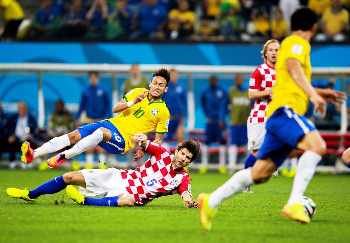 Foto mostrando um jogo de futebol do Brasil contra Croácia. Na imagem, Neymar está recebendo uma rasteira de um jogador do time adversário, enquanto seu companheiro toca a bola