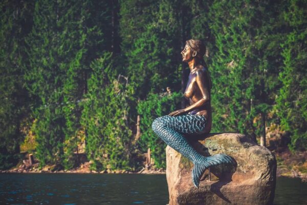 Estátua de uma sereia sentada em uma rocha no meio de um lago. O monumento parece ser de cobre e ressalta a causa azulada da criatura mitológica