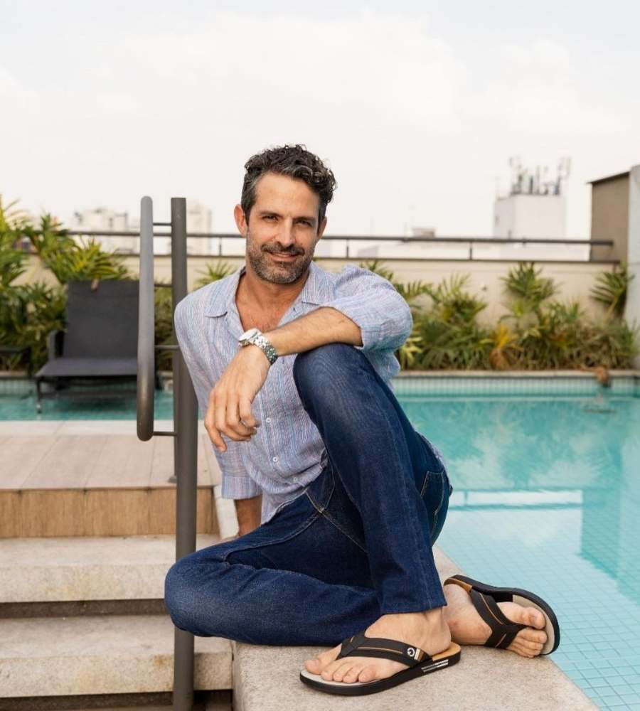 Foto do ator sentado, com o braço apoiado em seu joelho, em chão ao lado de piscina, área com escada, plantas e cadeiras. Ele usa calça jeans, camisa, sandália e relógio no pulso.