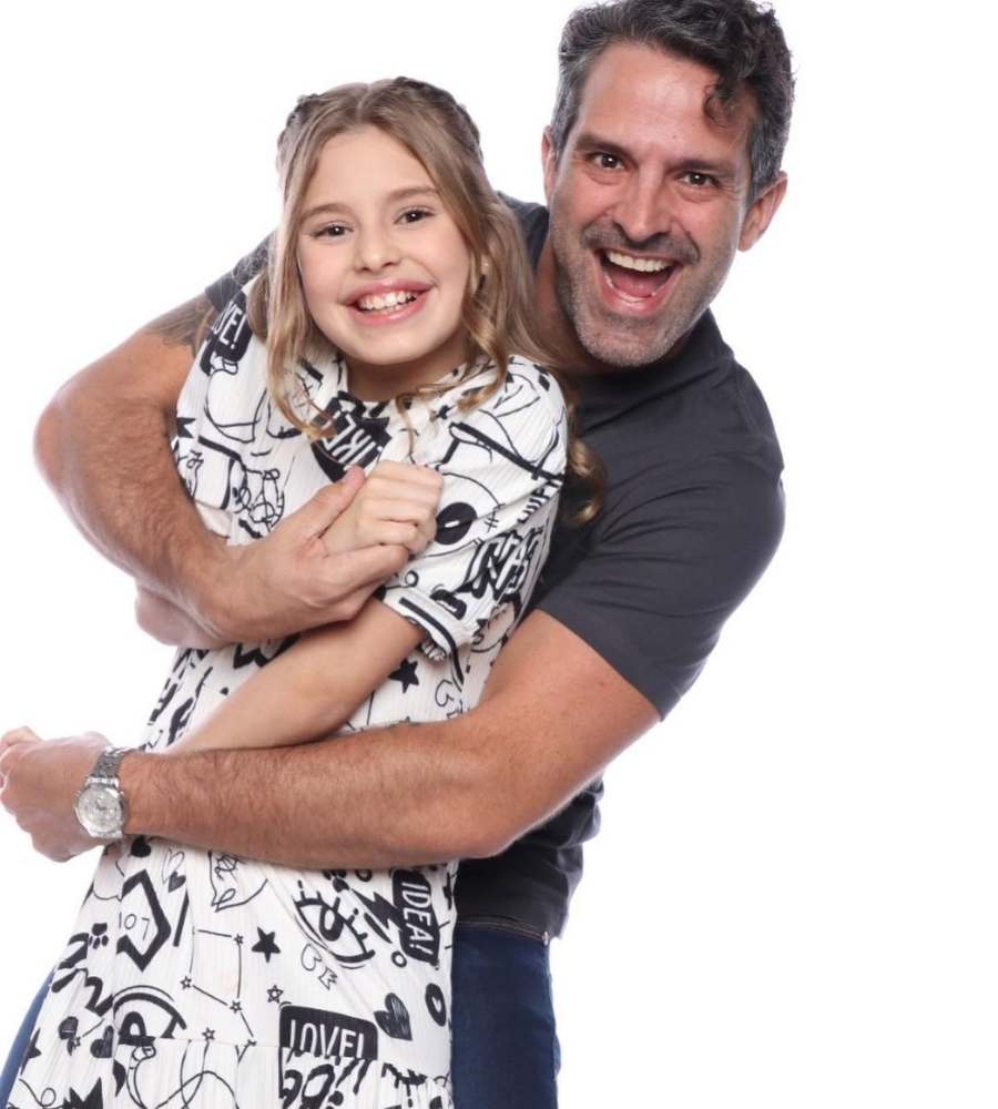 Foto de Iran e sua filha, Laura. Os dois estão sorridentes e abraçados. Ele usa camiseta cinza e ela vestido branco com desenhos pretos.