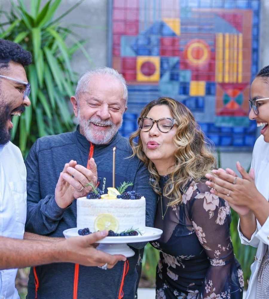 Foto de Lula sorridente batendo palma e Janja soprando velinha de bolo, segurado por um homem. Na foto tem também uma mulher que está batendo palma. Eles comemoram o 56º aniversário de Janja.