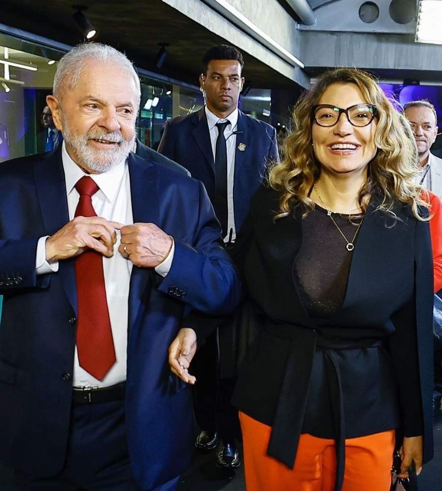 Foto de Lula e Janja juntos. Os dois estão sorridentes, dentro dos estúdios da Rede Bandeirantes. Atrás deles, tem dois homens.