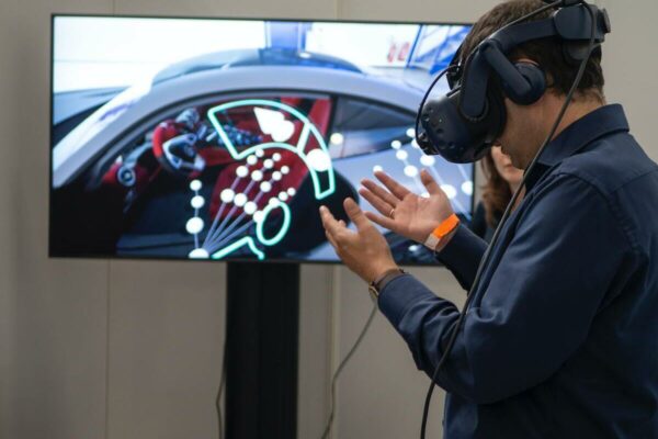 Homem com óculos de realidade virtual olhando para suas mãos e, na sua frente, há uma TV mostrando o que ele está vendo