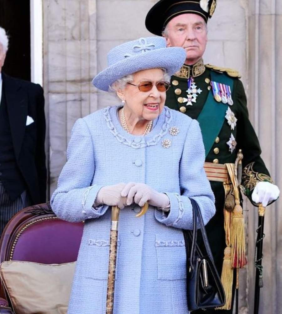 Foto da Rainha Elizabeth usando roupa azul bebê e segurando muleta. Ela está acompanha da soldados oficiais do Reino Unidos.