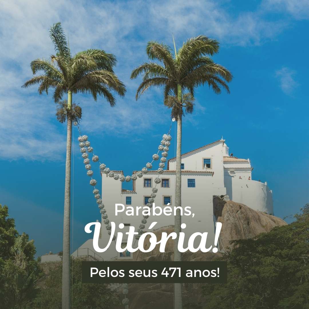 Foto da cidade de Vitória, imóvel branco com janelas, na frente tem mata com duas palmeiras. O céu está azul.