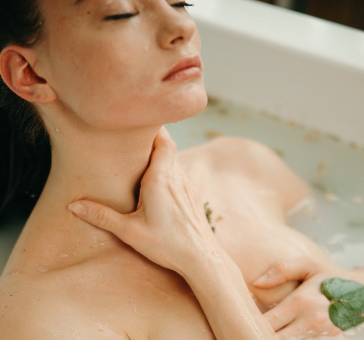 mulher branca de cabelos longos e escuros em uma banheira, tomando banho com uma folha verde que lembra um louro
