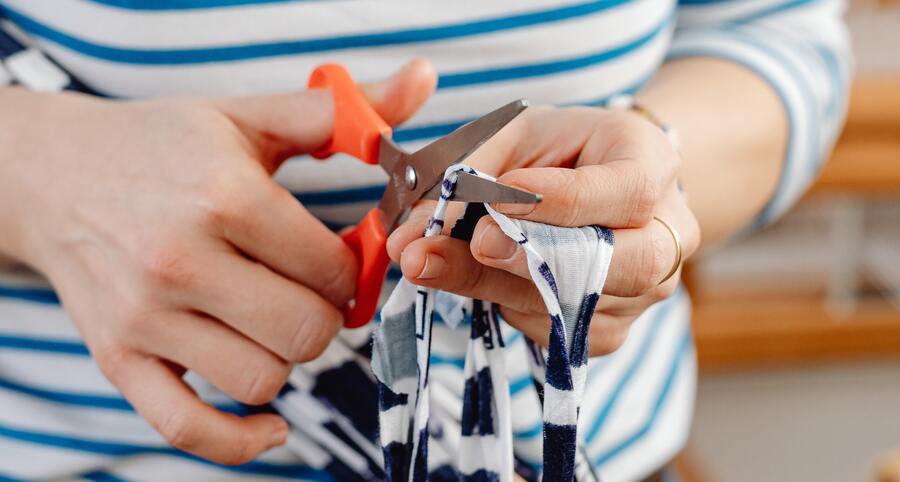 uma mão feminina com uma tesoura laranja cortando um pedaço de tecido branco e azul