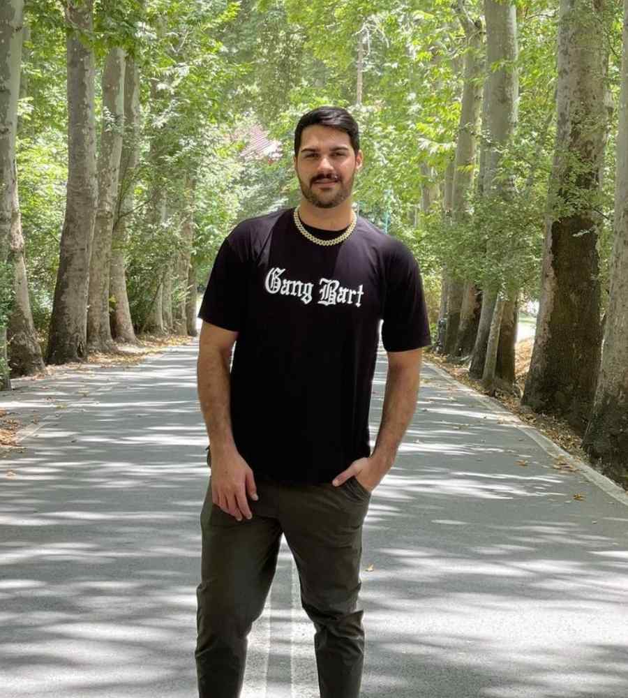 Foto do peão em rua com árvores dos dois lados. Ele está de pé no meio e usando camiseta preta, calça verde-militar e corrente dourada no pescoço.