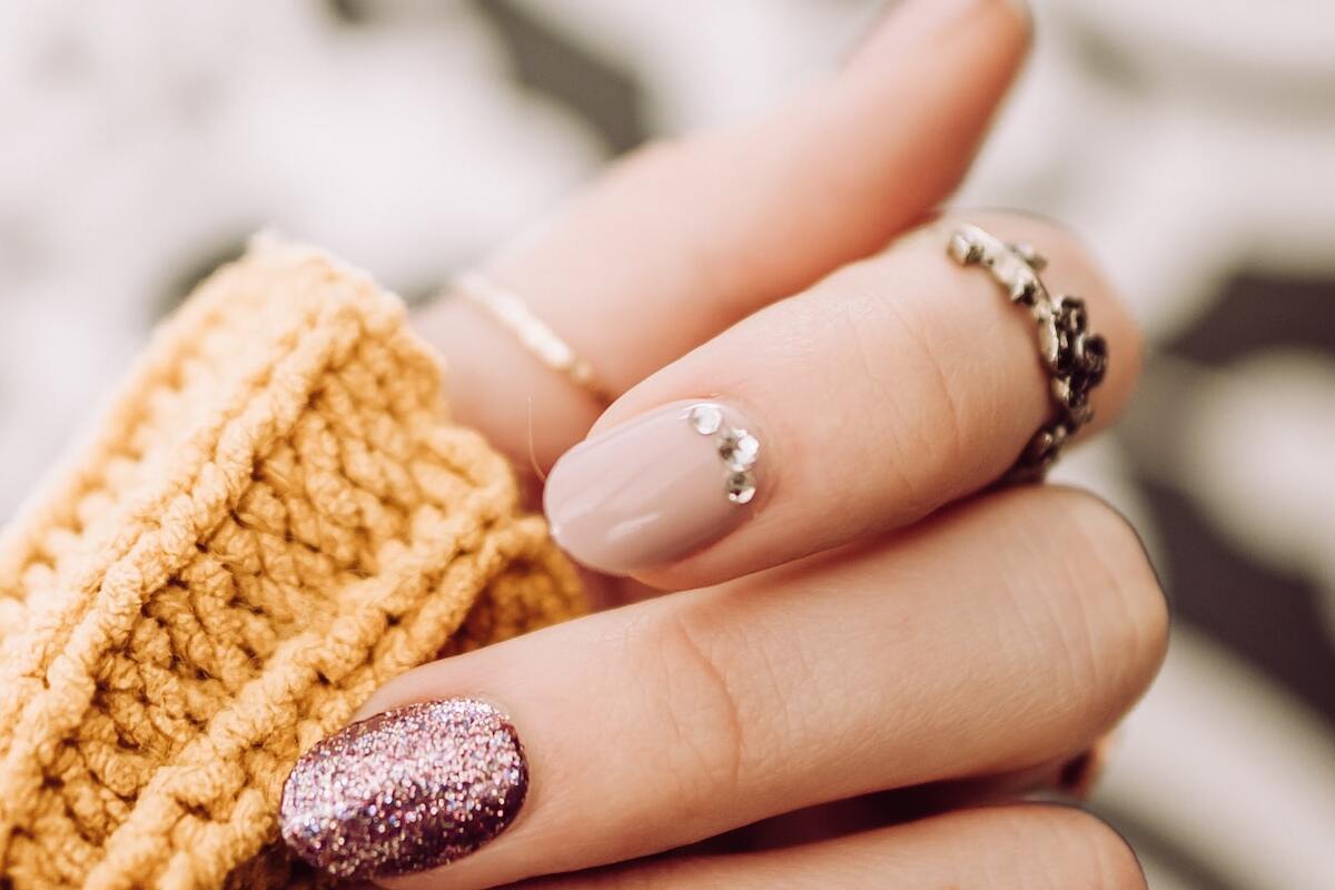 mão de uma mulher com vários anéis. Suas unhas estão pintadas de um tom metálico e uma delas possui pedrarias